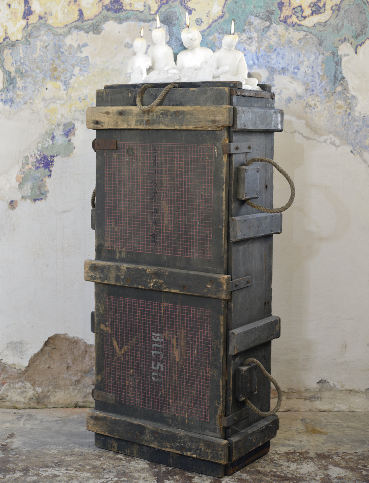 Four white Buddha figures with lit wicks standing on a dark wooden ammunition crate with rope handles and red grid panels